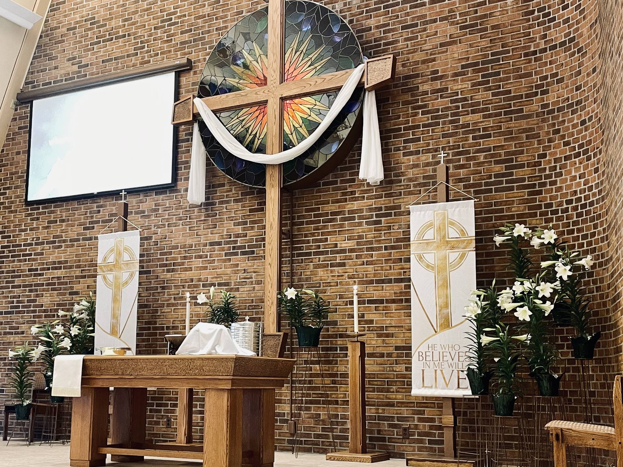 Bethany church altar with wooden cross, candles, and Easter banners reading 'He Who Believes Shall Live'